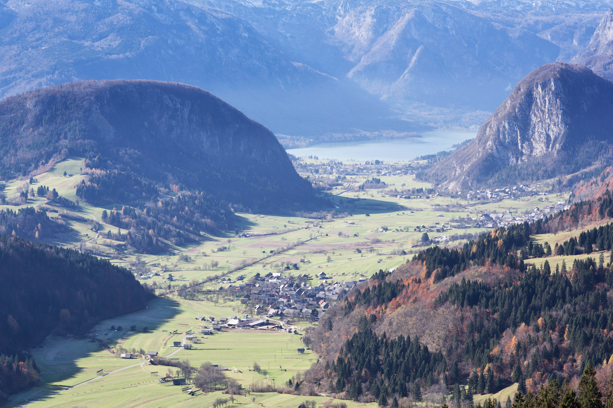 The photograph depicts a mountain valley with two settlements in the middle. A river runs through the valley. The hillsides are partly forested and partly covered with meadows, and a lake is visible in the distance.